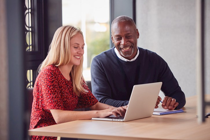 Woman and man, smiling, sitting at a desk and looking at a laptop.