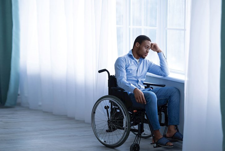 Man sitting in wheelchair by window with head on his hand, looking sad