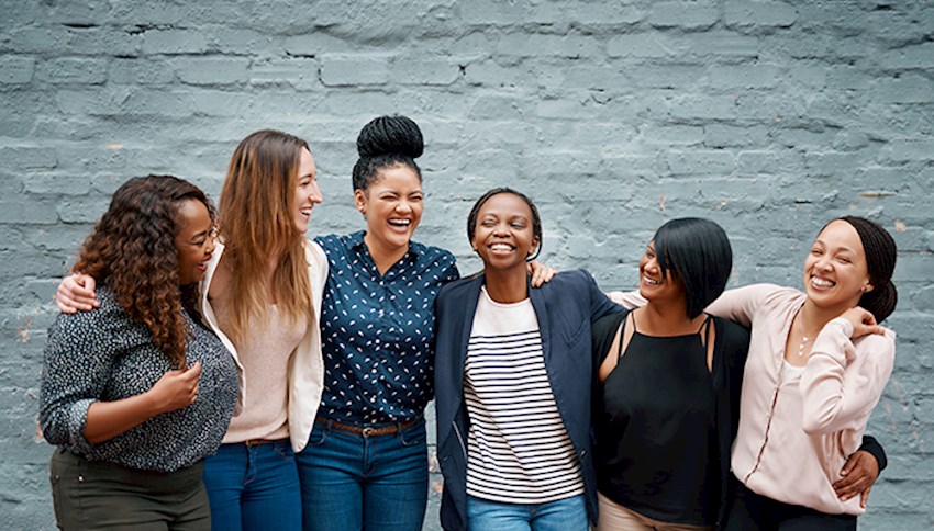 Diverse group of woman with arms around each other smiling in front of gray brick wall