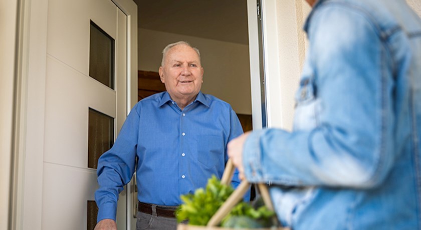 Senior man using cane standing in doorway greeting woman holding bag of food
