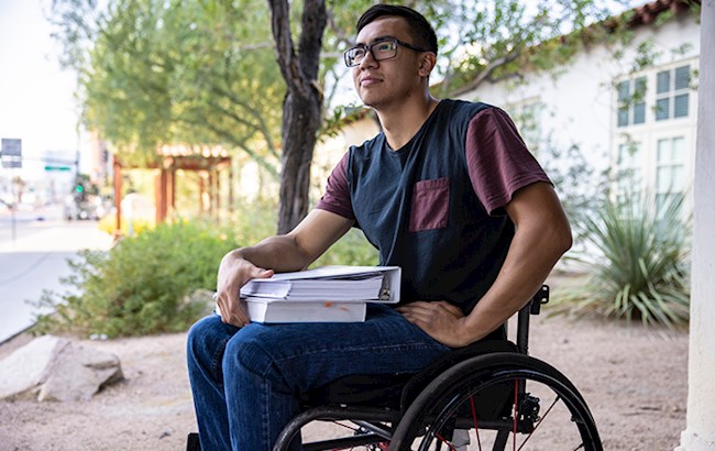 Male college student outside in a wheelchair holding a binder and a textbook