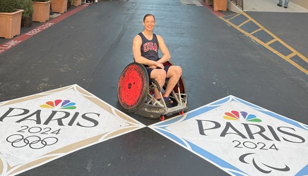 AOTA Member Sarah Adam, Paralympian female rugby player sits between two Paris Olympics flags