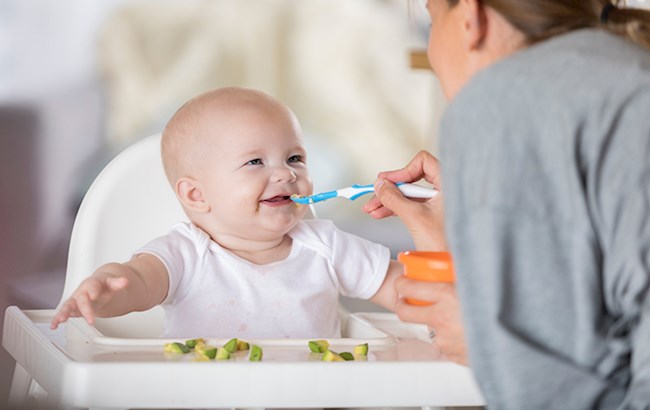 Baby in high chair smiling as woman feeds him with blue spoon
