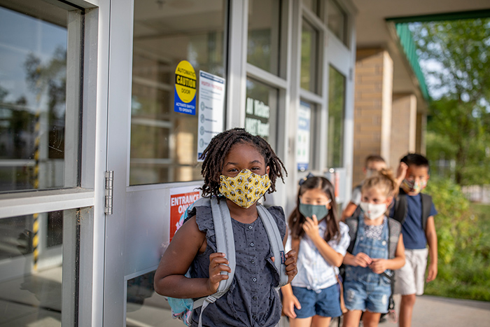 Young children wearing face masks wearing backpacks lined up outside of school building