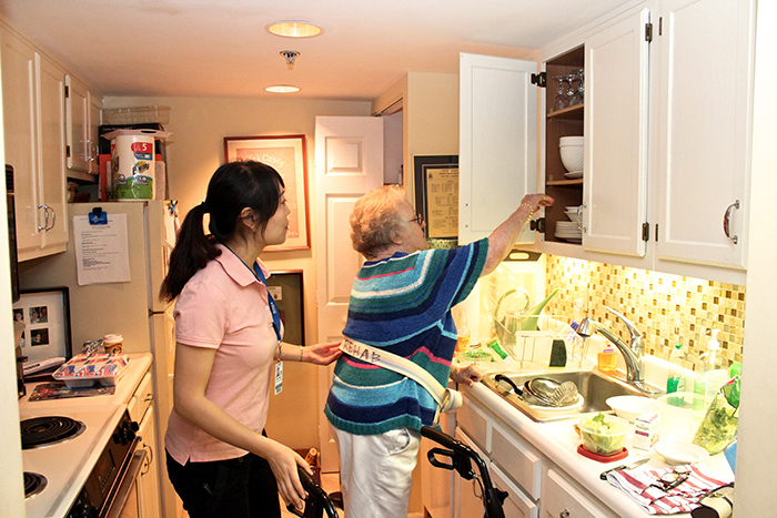 Senior woman in kitchen reaching upper cabinet with occupational therapist holding support belt