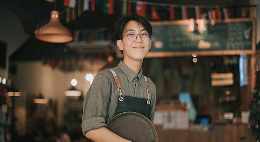 Smiling teenage boy wearing apron holding a round tray inside a restaurant