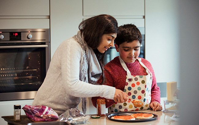 Woman in kitchen helping young boy wearing apron cook pizza
