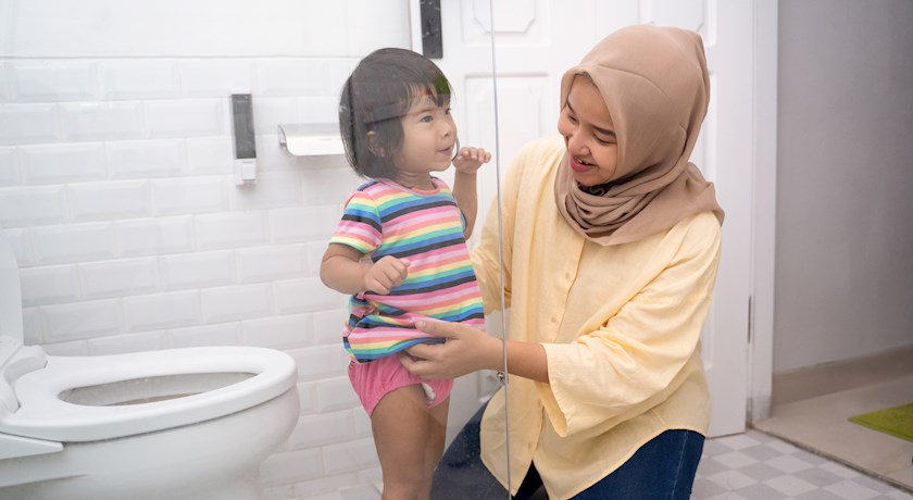 Toddler stands next to a toilet in a bathroom with parent while the parent fixes the toddler's clothes.