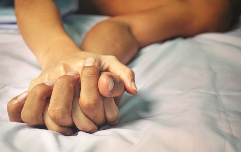 Arms on a bed from two people with hands grasping each other.