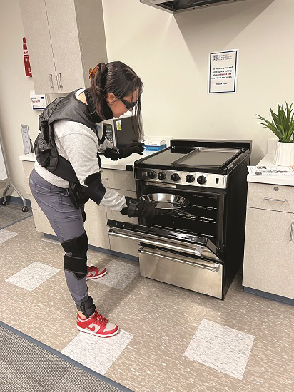 An OT student bending to place a tray in the oven, using geriatric simulation equipment to understand the physical limitations and challenges faced by elderly individuals during meal preparation tasks