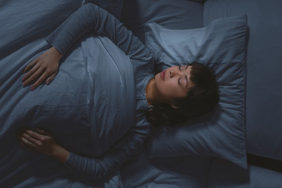 Woman with brown hair and long sleeve top, sleeping on gray sheets