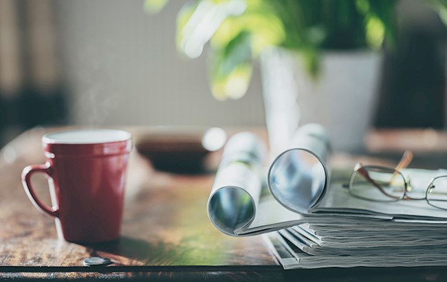 Desk with stack of magazines and a pair of glasses on top alongside a cup of coffee and a plant