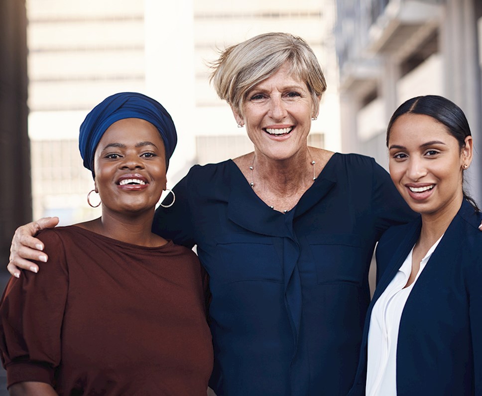Three multi ethnic woman professionally dressed and smiling at camera and middle woman has arms around the other two