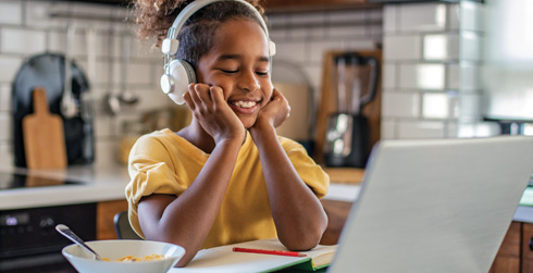 Smiling child sitting at kitchen counter with an open laptop, listening to headphones