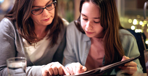 An adult and adolescent look at a book at a table while pointing at the pages.